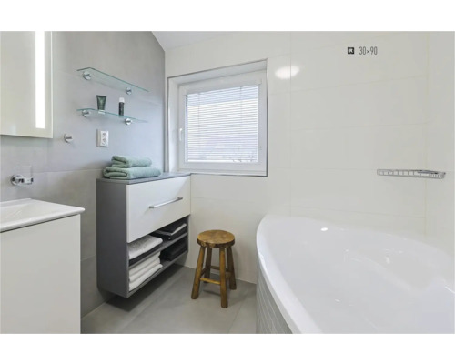 Salle de bains lumineuse avec baignoire, lavabo, meuble sous-vasque, serviettes, tabouret en bois et fenêtre avec store