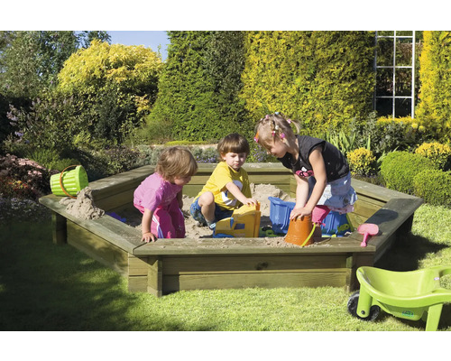 Trois enfants jouent avec des jouets dans un bac à sable hexagonal dans le jardin.