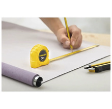 Scène avec une personne traçant une ligne au crayon sur une pièce, assistée par un mètre ruban Stanley de 3 mètres, avec un produit en rouleau et un cutter à proximité.