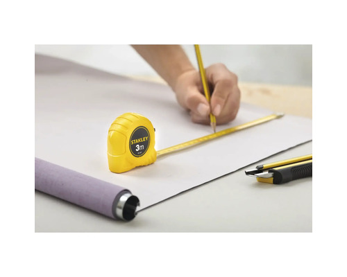 Scène avec une personne traçant une ligne au crayon sur une pièce, assistée par un mètre ruban Stanley de 3 mètres, avec un produit en rouleau et un cutter à proximité.