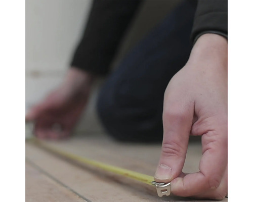 Une personne mesure un sol avec un mètre ruban jaune.