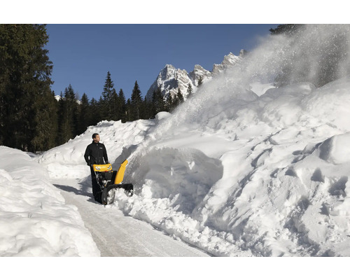 Eine Schneefräse räumt eine verschneite Straße frei, im Hintergrund sind Bäume und Berge zu sehen.