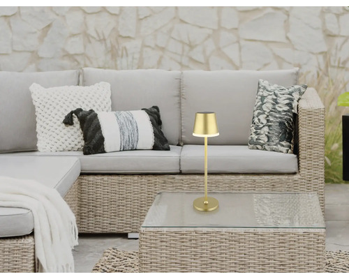 Goldfarbene Tischleuchte auf einem Gartentisch neben einer Rattancouch mit Kissen.