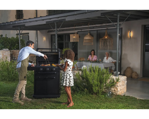 Szene im Freien mit einem Mann, der an einem Grill Speisen zubereitet, während zwei Frauen an einem Tisch sitzen