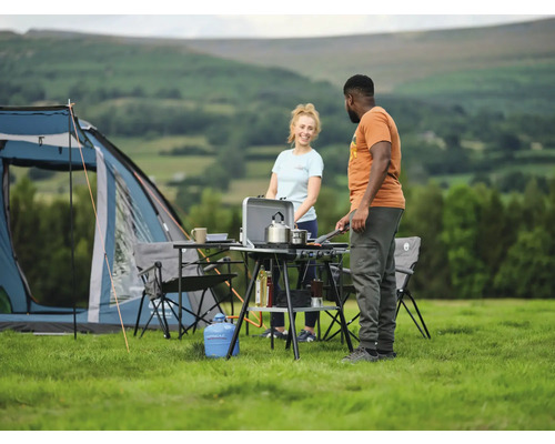 Scène de camping avec tente, table de camping, réchaud de camping et deux personnes cuisinant à l''extérieur.