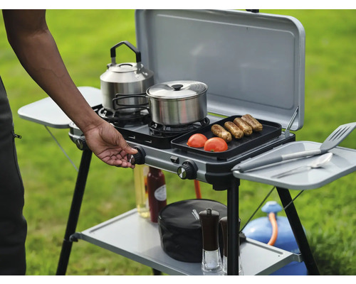 Réchaud de camping avec ustensiles de cuisine, aliments et cartouche de gaz sur une prairie.