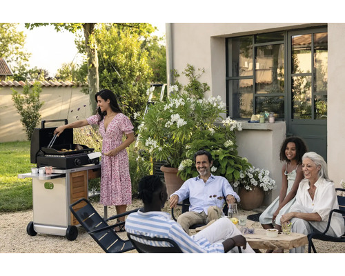 Scène de jardin avec une femme cuisinant sur un barbecue à gaz, tandis qu''un groupe de personnes est assis ensemble