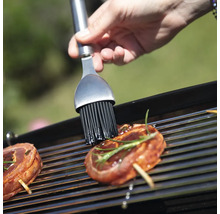 Eine Person bestreicht mit einem Pinsel Fleisch auf einem Grill.