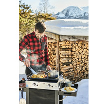 Un homme cuisine dans la neige sur un gril Eno devant une pile de bois et un paysage de montagne.