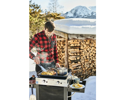 Un homme cuisine dans la neige sur un gril Eno devant une pile de bois et un paysage de montagne.