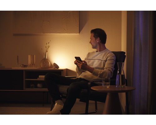 Un homme est assis avec un smartphone dans un fauteuil dans un salon avec un buffet, une lampe et une table.