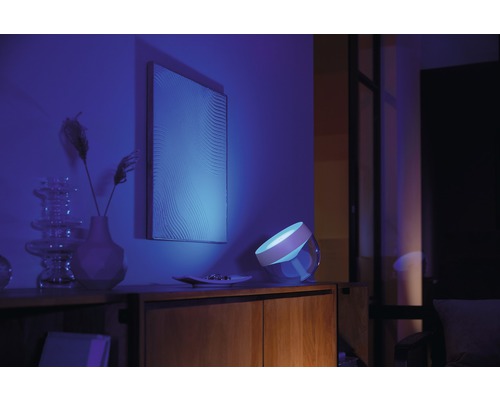 Scène avec une table de chevet sur laquelle se trouve une lampe émettant une lumière bleue, un tableau au mur et quelques éléments décoratifs.