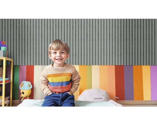 Un garçon souriant est assis dans une chambre d''enfant avec du papier peint rayé et une décoration murale colorée.