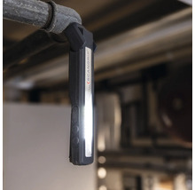 Scangrip Logo. Arbeitsleuchte mit Magnet und LED-Licht hängt an einem Rohr.