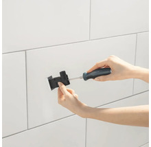 Eine Person befestigt eine schwarze Duschhalterung mit einem Schraubenzieher an einer weissen gefliesten Wand.