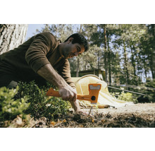 Ein Mann benutzt einen Hammer, um einen Hering in den Boden zu schlagen, mit einem Zelt im Hintergrund.