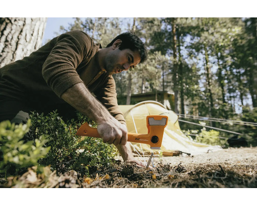 Ein Mann benutzt einen Hammer, um einen Hering in den Boden zu schlagen, mit einem Zelt im Hintergrund.