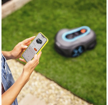 Une personne contrôle un robot tondeuse Gardena avec un smartphone dans le jardin.