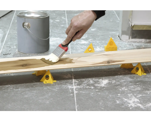 Holzbrett wird mit einem Pinsel gestrichen, Farbdose und Malereihilfen sind sichtbar.