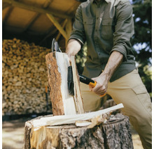 Une personne fend du bois avec une hache de marque Fiskars sur une souche d''arbre devant une pile de bois.