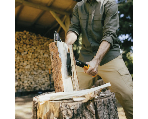 Une personne fend du bois avec une hache de marque Fiskars sur une souche d''arbre devant une pile de bois.