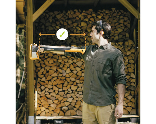 Un homme tient une hache à fendre Fiskars X25 dans sa main tendue devant une pile de bois. L''accent est mis sur la bonne longueur de bras.