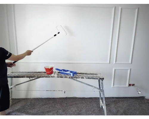 Un homme peint un mur avec un rouleau à peinture et une rallonge. Un seau de peinture et une grille à peinture sont posés sur un échafaudage.