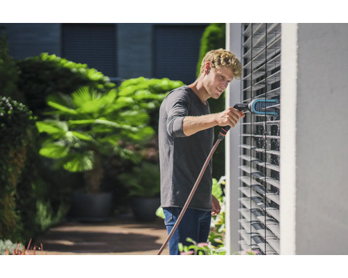 Mann reinigt Jalousien mit einer Bürste und einem Wasserschlauch im Garten.