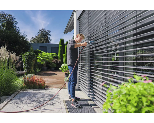 Mann reinigt Aussenjalousien mit einer Bürste und Wasserschlauch im Garten.