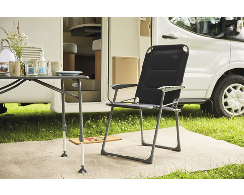 Szene mit einem Campingstuhl, einem Campingtisch, Geschirr und einem Wohnmobil im Freien.