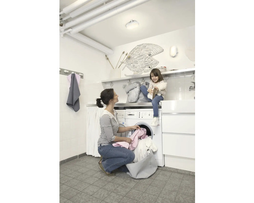 Frau befüllt eine Waschmaschine mit Wäsche, während ein Kind auf der Maschine sitzt