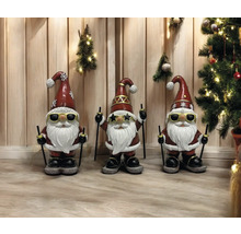 Trois nains de jardin avec des lunettes de soleil, des skis et des bonnets de Noël se tiennent devant un mur en bois décoré de décorations de Noël.