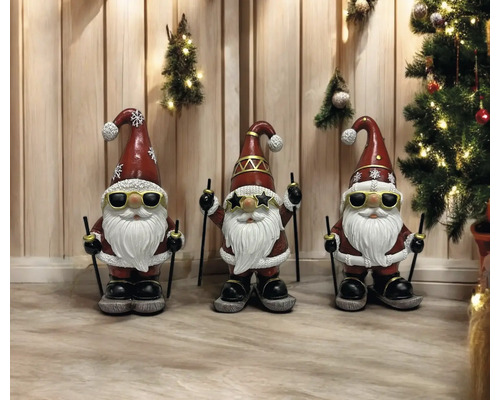 Drei Gartenzwerge mit Sonnenbrille, Skiern und Weihnachtsmützen stehen vor einer Holzwand mit Weihnachtsschmuck.