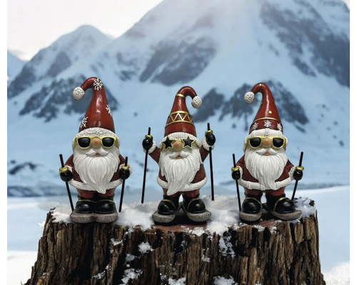 Drei Gartenzwerge mit Sonnenbrillen und Skiern auf einem Baumstumpf vor einem Bergpanorama