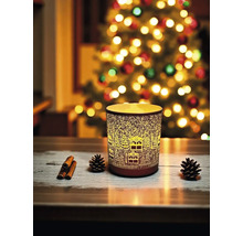 Dekorative Kerze mit Winterdorf-Design neben Tannenzapfen und Zimtstangen vor einem Weihnachtsbaum.