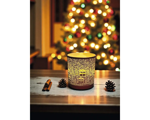 Dekorative Kerze mit Winterdorf-Design neben Tannenzapfen und Zimtstangen vor einem Weihnachtsbaum.