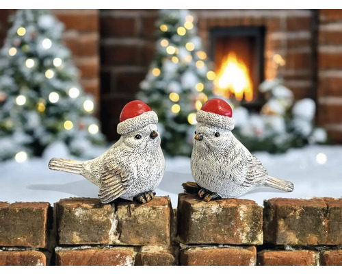 Deux oiseaux décoratifs avec des bonnets de Noël sur un mur de briques devant une cheminée et des sapins de Noël