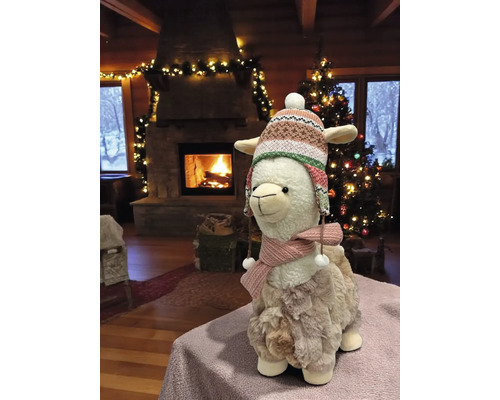 Dekoratives Lama mit Mütze und Schal vor Kamin und Weihnachtsbaum.