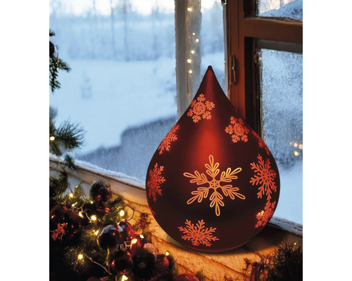 Dekorative, rote Schneeflockenlampe auf einer Fensterbank mit verschneiter Landschaft im Hintergrund.