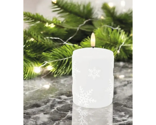 Bougie décorative à motif de flocons de neige devant des branches de sapin et des lumières.