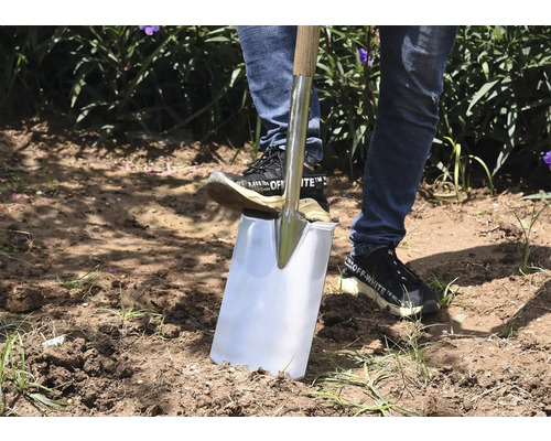 Eine Person benutzt einen Spaten mit Holzgriff im Garten, um Erde auszuheben.