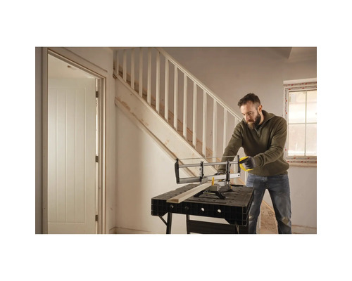 Un homme scie du bois avec une scie à main et une boîte à onglets sur un établi dans une pièce avec un escalier et une porte.