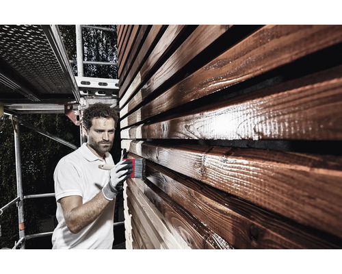 Un homme peint une façade en bois avec un pinceau et une lasure de protection du bois depuis un échafaudage