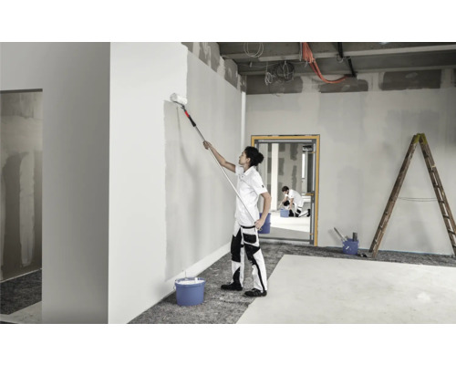 Une personne peint un mur dans une pièce avec un rouleau à peinture sur une tige télescopique. Une autre personne et une échelle sont visibles en arrière-plan.