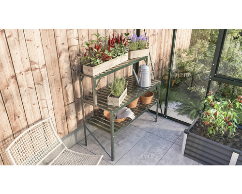 Gartenszene mit einem Pflanztisch aus Metall, bepflanzten Holzkästen, einer Giesskanne und Gartengeräten in einem Gewächshaus