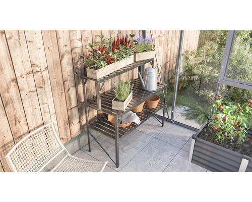 Drei Etagen Pflanztisch aus Metall mit Pflanzen, Töpfen, Giesskanne und Gartenhandschuhen auf einem Balkon neben einem Gewächshaus.