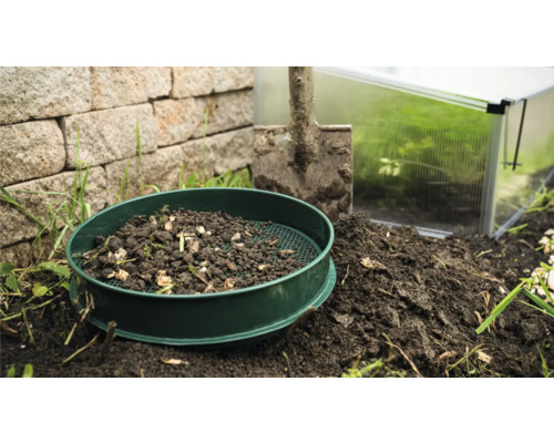 Gartensieb mit Erde, Schaufel und Gewächshaus im Hintergrund