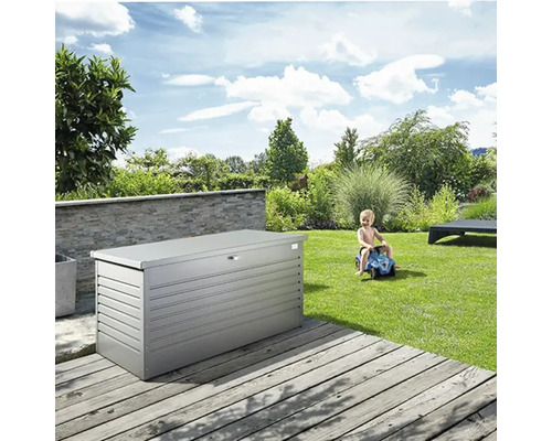 Coffre de jardin gris sur une terrasse en bois, avec un enfant jouant et un paysage de jardin verdoyant en arrière-plan
