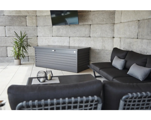 Espace extérieur avec un coffre de rangement gris, un canapé, une table et une plante.
