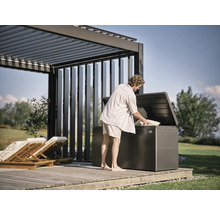 Mann befüllt eine Gartenbox unter einer Pergola auf einer Holzterrasse mit Kissen.